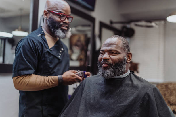 A mature adult black man smiles at his reflection with satisfaction after receiving a hair cut and beard trim at his local barbershop.